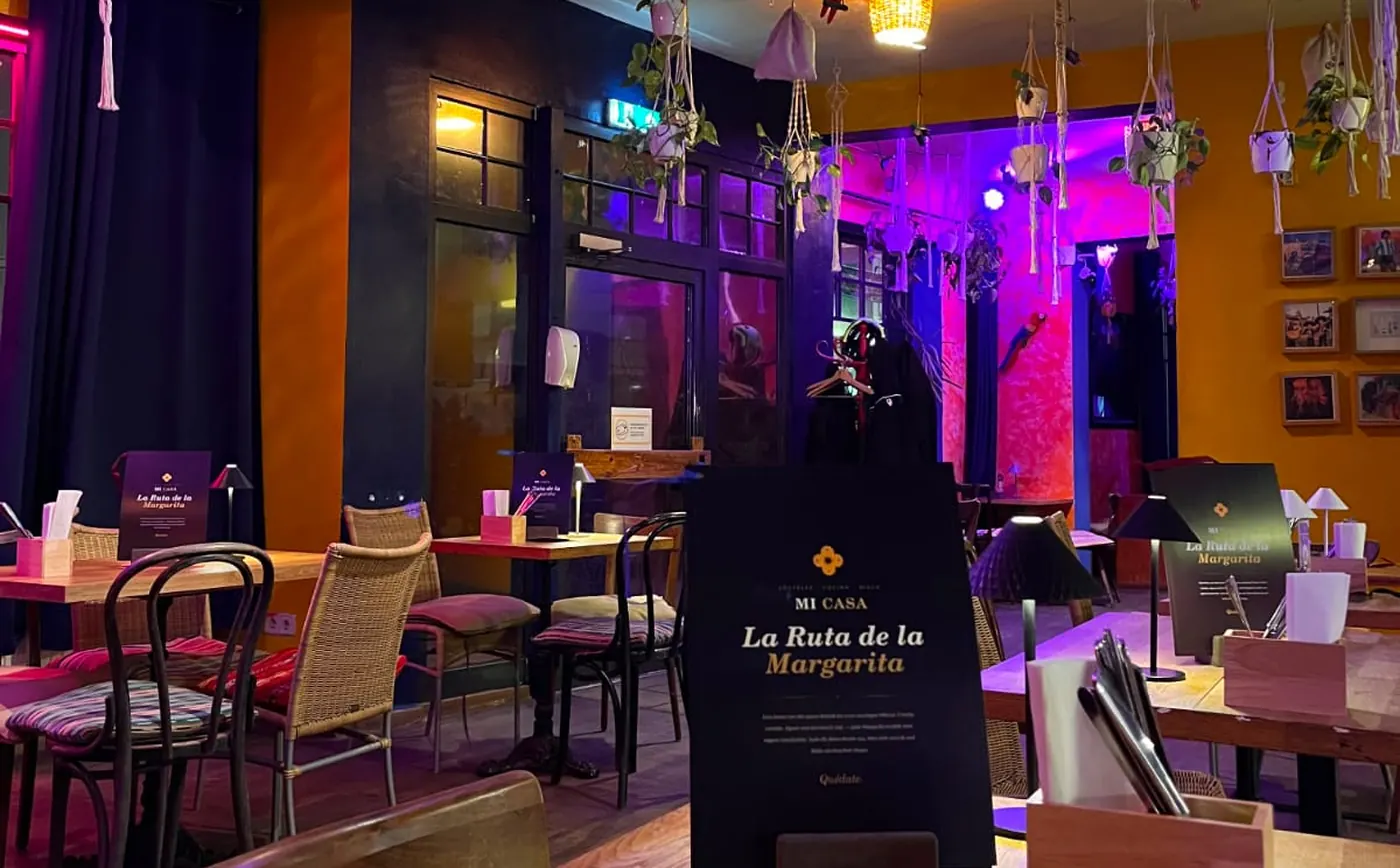 Mi Casa am Lend innen – Gastraum mit Hängepflanzen, warmem Licht und der Karte „La Ruta de la Margarita“ am Tisch